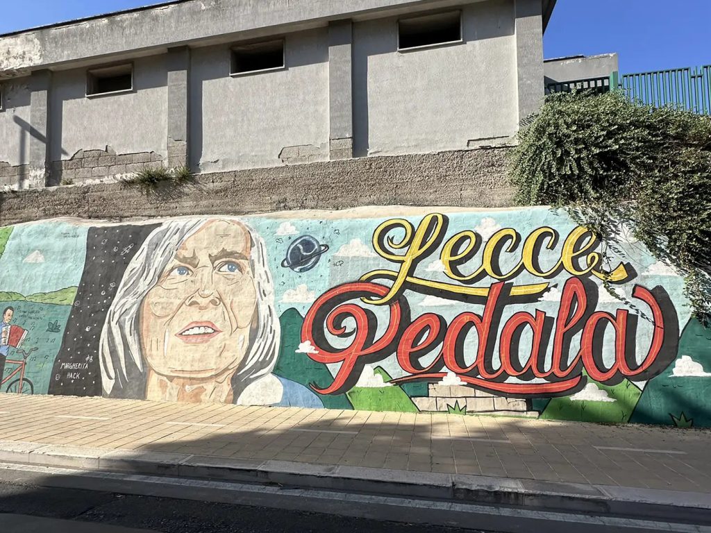 Murale con il volto di una donna anziana e la scritta Lecce Pedala su un muro cittadino