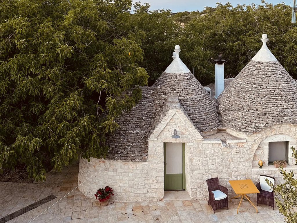 Trullo in pietra con tetti a cono e tavolino con sedie all'esterno tra la vegetazione in Puglia