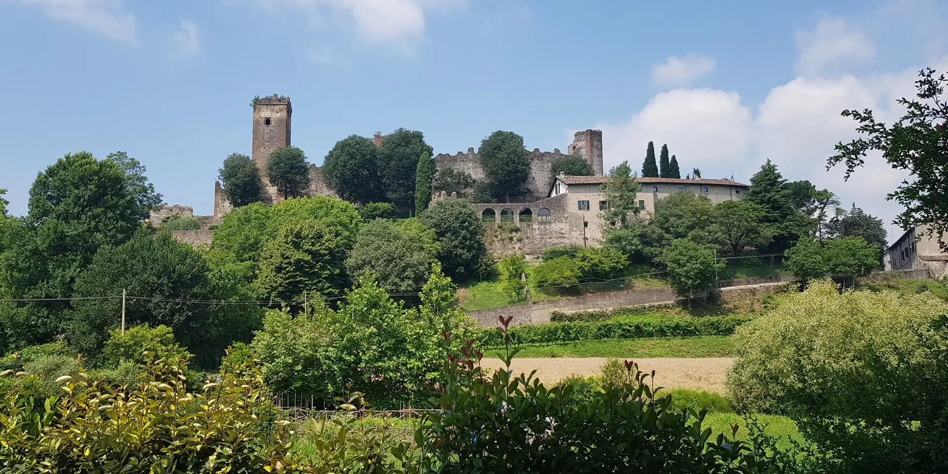 Antico borgo con torre e mura circondato da vegetazione sotto un cielo con nuvole bianche