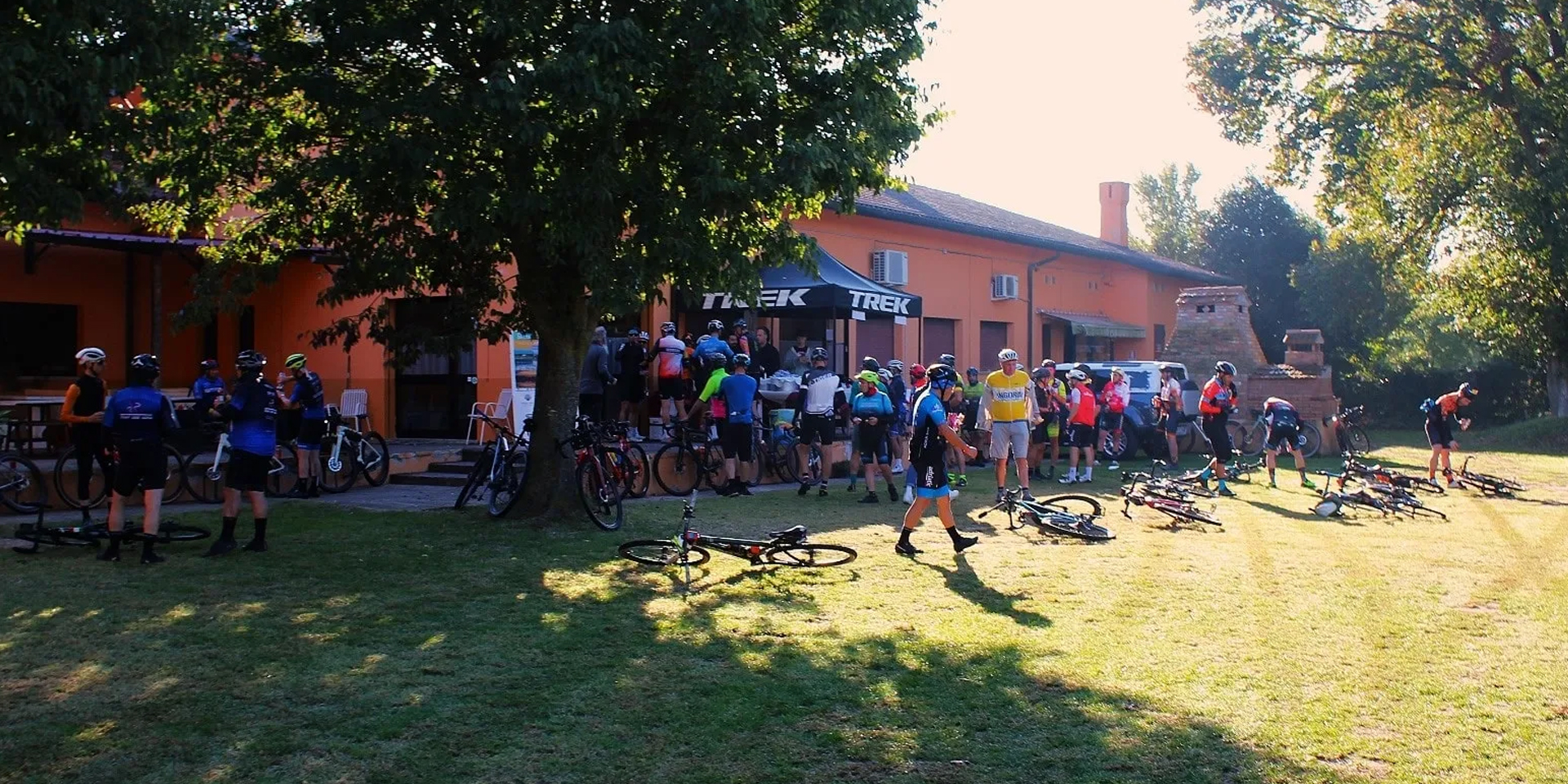 Partecipanti a una pedalata riuniti all'ombra di un grande albero davanti a una casa rossa nel Delta del Po