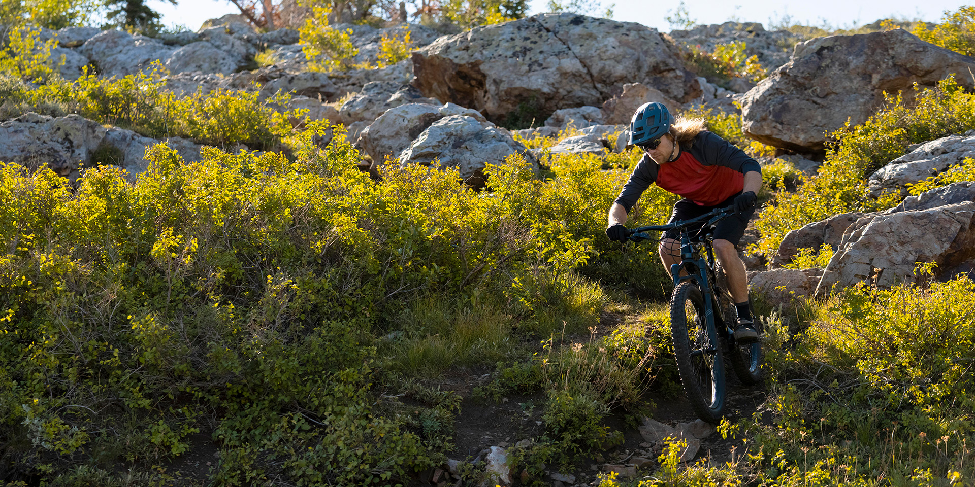 Rider in MTB affronta un single track tra arbusti verdi e massi illuminati dal sole